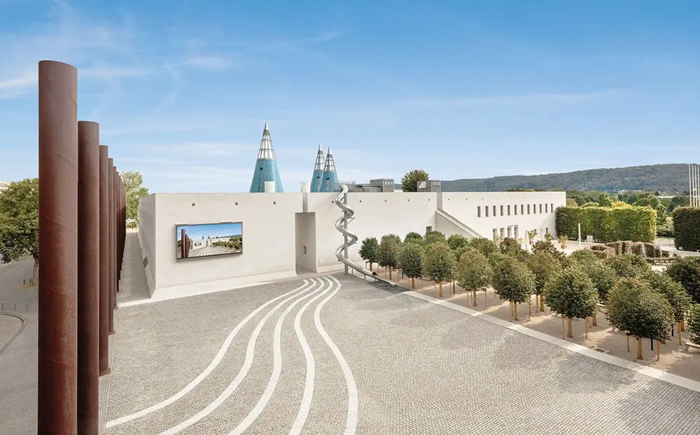 Blick auf den gepflasterten Vorhof der Bundeskunsthalle. Zur rechten stehen Bäume in einer Allee, die Bundeskunsthalle befindet sich im hinteren drittel mit ihrer Rutsche vom Dachgarten und den drei Spitzdächern.